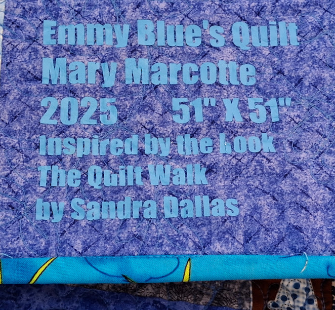 Photo of a quilt label that reads, Emmy Blue's Quilt; Mary Marcotte; 2025; 51" X 51"; Inspired by the book The Quilt Walk by Sandra Dallas